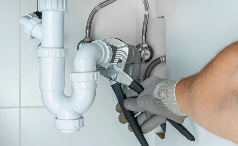 Plombier professionnel en tenue de travail intervenant en urgence dans une salle de bain, inspectant une fuite d'eau ou un problème de tuyauterie sous un lavabo. Le plombier, équipé d'outils professionnels, effectue un diagnostic. La scène capture l'action et la disponibilité immédiate du professionnel face à une situation d'urgence domestique. Éclairage naturel, environnement réaliste d'intérieur d'habitation.